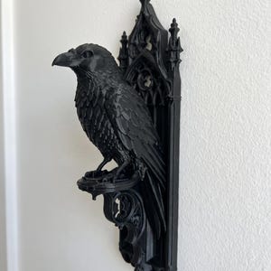 May include: A matte black wall-mounted sculpture featuring a detailed raven perched on a decorative bracket. The raven is positioned in front of a vertical panel with intricate pointed arch and tracery patterns reminiscent of gothic architecture.