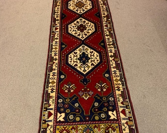 Op de afbeelding: Een lang, smal rood tapijt met een geometrisch patroon in tinten blauw, crème en goud. Het tapijt heeft een franje rand.