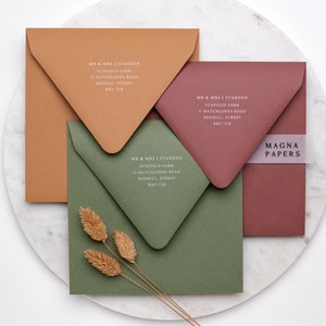 May include: Three square envelopes in different colors: brown, green, and red. The envelopes are all the same size and have a flap on the top. The envelopes are all addressed with the same text: "MR & MRS J STANDEN, SUNFIELD FARM, II HATCHLANDS ROAD, REDHILL, SURREY, RHS 7UB". The text is in a white font. A white ribbon with the text "MAGNA PAPERS" is on the red envelope.
