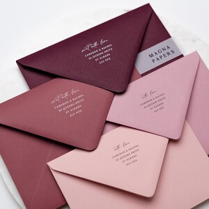May include: Four burgundy and pink envelopes with white calligraphy on the front. The envelopes are stacked on top of each other. The top envelope has the text "sent with love" and the address "CAMERON & RACHEL, 30 QUEENS DRIVE, ST ALBANS, ALS 400". The second envelope has the text "sent with love" and the address "CAMERON & RACHEL, 30 QUEENS DRIVE, ST ALBANS, ALS 400". The third envelope has the text "with love" and the address "CAMERON & RACHEL, 30 QUEENS DRIVE, ST ALBANS, ALS 400". The fourth envelope has the text "with love" and the address "CAMERON & RACHEL, 30 QUEENS DRIVE, ST ALBANS, ALS 400". The envelopes are on a white surface. The text "MAGNA PAPERS" is on a white label on the top envelope.