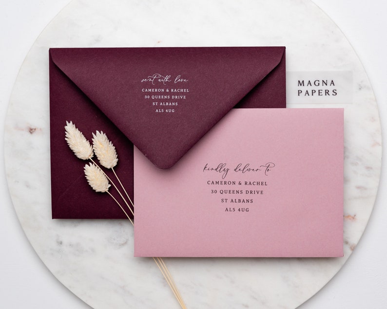 May include: Two envelopes, one burgundy and one pink, with the text "sent with love" and "kindly deliver to" on them. The envelopes are addressed to Cameron & Rachel, 30 Queens Drive, St Albans, AL5 4UG. A clear sticker with the text "MAGNA PAPERS" is on the pink envelope.
