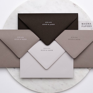 May include: Three envelopes in different shades of brown with the text "With Love Alice & Jason" printed on each. The envelopes are stacked on a white marble surface. The text "MAGNA PAPERS" is printed on a clear sticker on the top envelope.