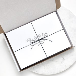 Puede incluir: Caja de regalo blanca abierta, atada con una cinta negra. La caja tiene un interior blanco. El texto "Father Of The Bride" est&aacute; impreso en la caja, junto con una direcci&oacute;n.