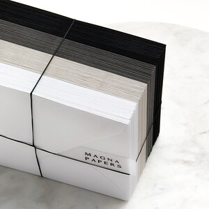 May include: A stack of white, grey, and black envelopes tied together with a black elastic band. The envelopes are labelled "MAGNA PAPERS".