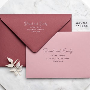 May include: Two pink and burgundy envelopes with the text "Daniel and Emily" and their address, "24 Earl Drive, Congleton, Cheshire, CW12 4AB". The envelopes are on a white marble surface. The text "MAGNA PAPERS" is on a clear sticker on the top right corner of the image.