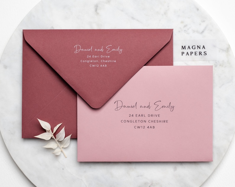 May include: Two pink and burgundy envelopes with the text "Daniel and Emily" and their address, "24 Earl Drive, Congleton, Cheshire, CW12 4AB". The envelopes are on a white marble surface. The text "MAGNA PAPERS" is on a clear sticker on the top right corner of the image.