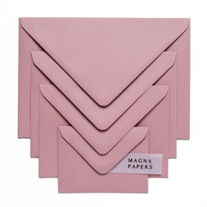 May include: A set of five dusty rose envelopes of varying sizes, nested inside each other. The smallest envelope has a white label with the text "MAGNA PAPERS" printed in black.