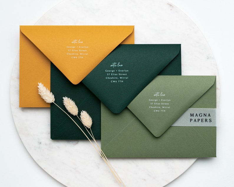 May include: Three envelopes in different colors: mustard yellow, forest green, and olive green. Each envelope has a white handwritten address label with the text "with love" and a name and address. The bottom envelope has a clear sticker with the text "MAGNA PAPERS" in black.