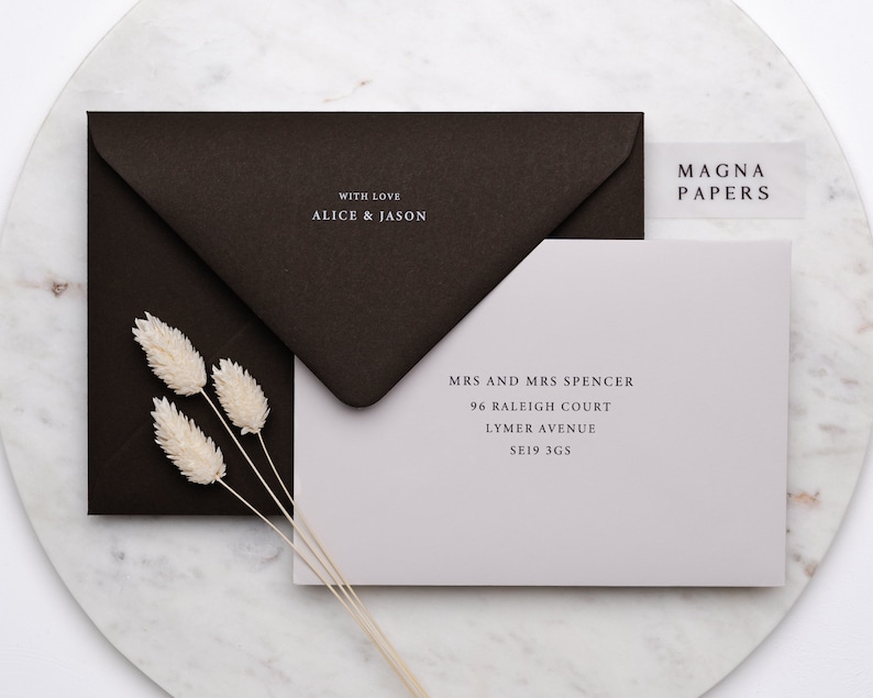 May include: A light grey envelope with a white address label that reads "MRS AND MRS SPENCER 96 RALEIGH COURT LYMER AVENUE SE19 3GS". The envelope is sitting on a white marble surface with a brown envelope and dried flowers.