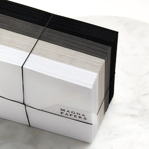 May include: A stack of white, gray, and black envelopes tied together with a black elastic band. The envelopes are labeled "MAGNA PAPERS".