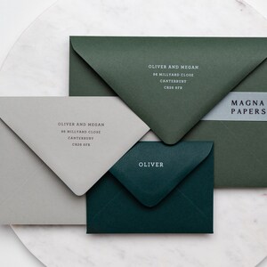 May include: Three envelopes in different colors: gray, dark green, and a lighter shade of green. The envelopes are addressed with the name "Oliver and Megan" and the address "96 Millyard Close, Canterbury, CR26 6FR". The lighter green envelope has the text "Oliver" on it. The dark green envelope has a white label with the text "MAGNA PAPERS" on it.