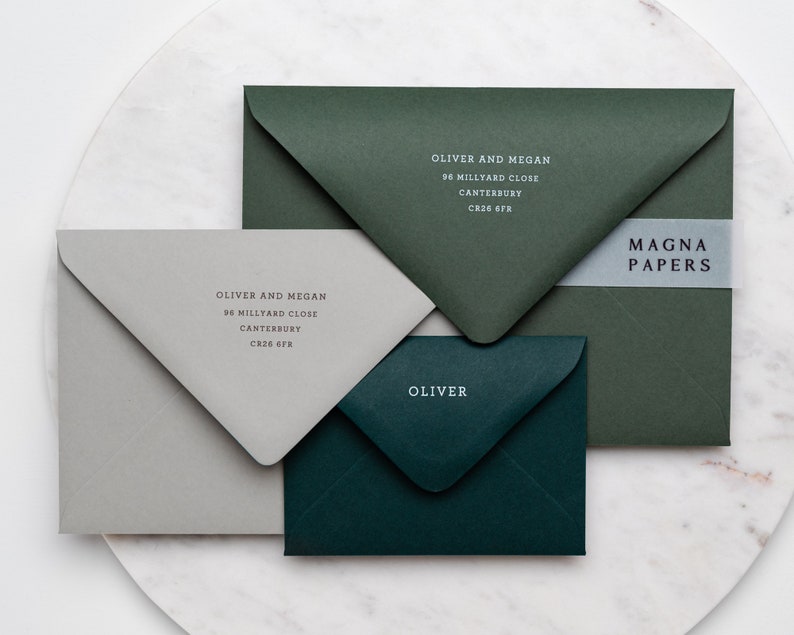 May include: Three envelopes in different colors: gray, dark green, and a lighter shade of green. The envelopes are addressed with the name "Oliver and Megan" and the address "96 Millyard Close, Canterbury, CR26 6FR". The lighter green envelope has the text "Oliver" on it. The dark green envelope has a white label with the text "MAGNA PAPERS" on it.