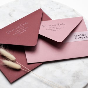 May include: Two burgundy and pink envelopes with white calligraphy. The envelopes are addressed to "Daniel and Emily" with a street address and postcode. The pink envelope has a clear sticker with the text "MAGNA PAPERS" printed in black.