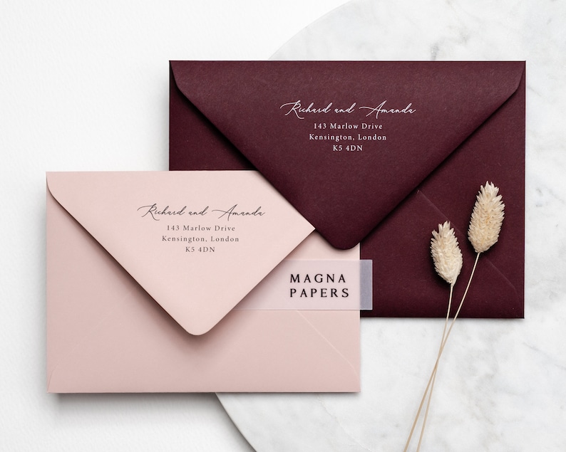 May include: Two envelopes, one burgundy and one blush pink, with the text "Richard and Amanda" and their address printed on them. The burgundy envelope has a white label with the text "MAGNA PAPERS" on it.