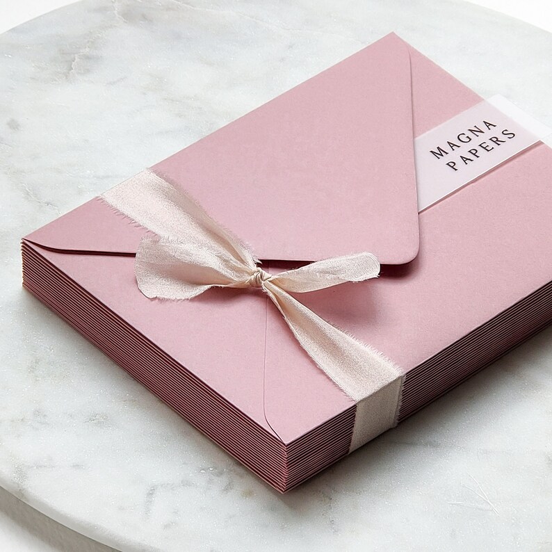 May include: A stack of blush pink envelopes tied with a blush pink ribbon. The envelopes have a clear sticker with the text "MAGNA PAPERS" on the top.