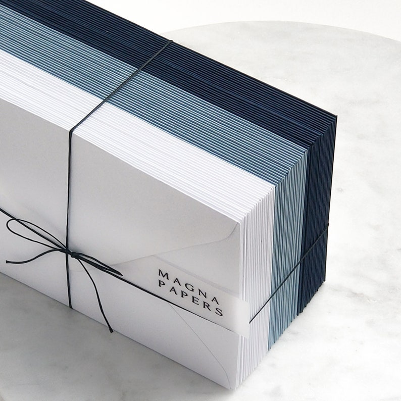 May include: A stack of white envelopes tied with black string. The envelopes are layered with blue and navy blue paper. The envelopes have the text "MAGNA PAPERS" printed on them.