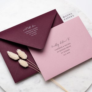 May include: Two envelopes, one burgundy and one pink, with the text "sent with love" and "kindly deliver to" on them. The text includes the names Cameron & Rachel, the address 30 Queens Drive, St Albans, and the postcode AL3 4UG. The envelopes are on a white surface with a decorative design.