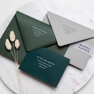 May include: Three green envelopes with white text on them. The text reads "Oliver and Megan" followed by an address. The envelopes are on a gray marble surface. A clear sticker with the text "MAGNA PAPERS" is on the top envelope.