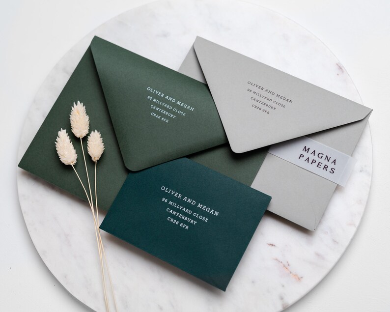May include: Three green envelopes with white text on them. The text reads "Oliver and Megan" followed by an address. The envelopes are on a gray marble surface. A clear sticker with the text "MAGNA PAPERS" is on the top envelope.