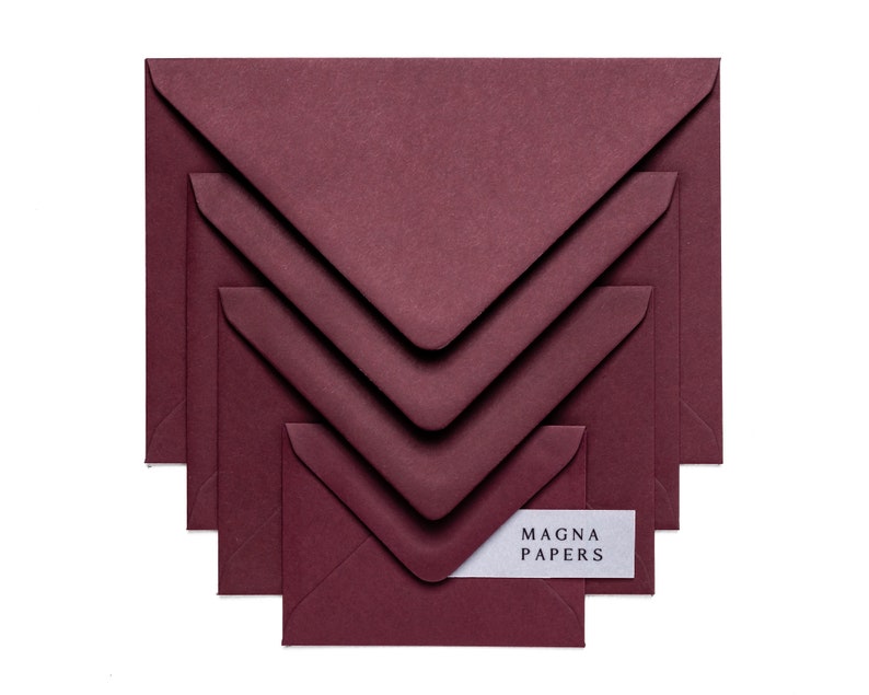 May include: Set of five burgundy envelopes of different sizes, stacked on top of each other. The smallest envelope has a white label with the text "MAGNA PAPERS" printed on it.