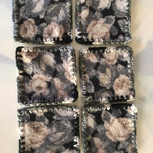 May include: Six square coasters with a floral pattern in shades of gray, black, and beige. Each coaster is edged with a dark gray stitch and a white stitch. The coasters are made of a soft, textured material.