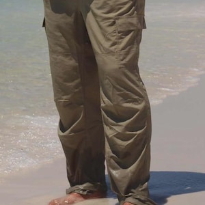 May include: Khaki cargo pants with multiple pockets. The pants are worn with a white button-down shirt. The person is standing on a sandy beach with their feet in the water.