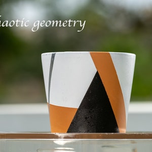 May include: A white ceramic planter with geometric black and orange designs. The planter is sitting on a clear glass surface.
