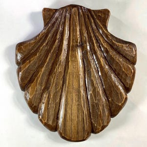 Può includere: Una conchiglia di legno intagliato con una superficie dettagliata e strutturata. Il guscio è di colore marrone caldo, con venature del legno visibili e creste intagliate. Il design ricorda l'arredamento costiero.