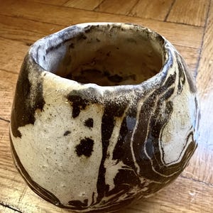May include: A small, rounded ceramic vessel with an irregular, hand-formed rim. The exterior features a marbled pattern of dark brown and off-white glaze with a visible crackle texture throughout the surface, creating a distinct swirling effect.