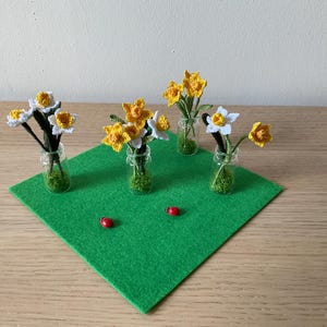 May include: A display of miniature crochet daffodils in small glass bottles, arranged on a green felt square. The daffodils have yellow and white petals. Two small red ladybugs are also on the green felt.