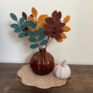 May include: A decorative arrangement featuring a brown glass vase with crocheted leaves in teal, mustard, and brown. A small white pumpkin and a woven placemat complete the fall-themed display.