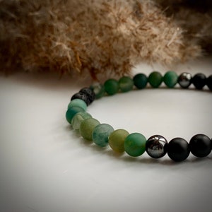 May include: A green and black beaded bracelet with a gradient of green beads. The bracelet is made with agate, lava rock, and hematite beads.