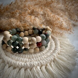 May include: A multi-layered bracelet made of green, white, and red beads. The bracelet is on a white crocheted surface with fringe.