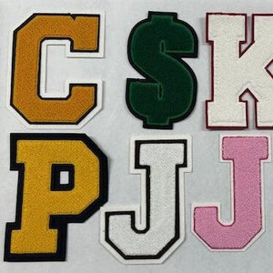 以下が含まれることがあります： 黄色、緑、白、ピンク、青など、さまざまな色のフェルト製の文字パッチが8つあります。文字は「C$KCPJJB」と綴られています。