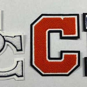 以下が含まれることがあります： 白、オレンジ、青のフェルト文字で「SCB」と綴られています。