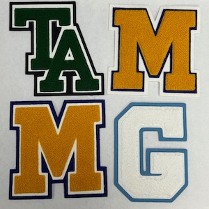 以下が含まれることがあります： 緑、金、金、青のフェルト文字「T」「A」「M」「G」。白いアウトラインが付いています。文字は白い背景に縫い付けられています。