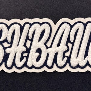 May include: A custom embroidered patch featuring the name KISHBAUGH in white, raised, cursive lettering with a dark navy blue outline and a thick white border, designed for application onto fabric surfaces.