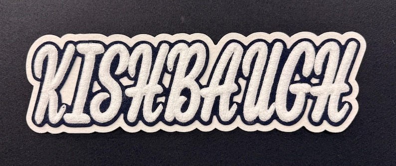 May include: A custom embroidered patch featuring the name KISHBAUGH in white, raised, cursive lettering with a dark navy blue outline and a thick white border, designed for application onto fabric surfaces.