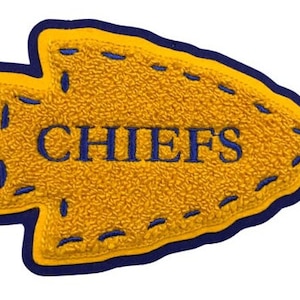 May include: A yellow and blue arrow-shaped patch with the word "CHIEFS" embroidered in blue. The patch has a textured surface and a blue border with decorative stitching.