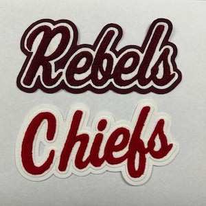 以下が含まれることがあります： 赤い文字で書かれた白いフェルトのパッチが2つ。上のパッチには「Rebels」と書いてあり、下のパッチには「Chiefs」と書いてあります。