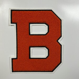 以下が含まれることがあります： 白の縁と黒のトリムが付いたオレンジ色の文字「B」のパッチ。文字は、起毛フェルトのような質感があります。パッチは、ジャケットやその他のアパレルのためである可能性があります。