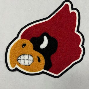 Puede incluir: Una aplicación de fieltro rojo, negro y amarillo de la cabeza de un cardenal con un pico blanco y una sonrisa de dientes.