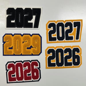 以下が含まれることがあります： 2026年、2027年、2029年の刺繍パッチのコレクション。パッチは、黒、金、赤、ネイビーブルーなど、さまざまな色の組み合わせを持つ、大学の文字スタイルです。