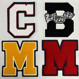 以下が含まれることがあります： 異なる色とスタイルのフェルトレターパッチ4つ。文字は「CMBM」と綴られています。「C」は赤色のアウトラインが付いた白、「B」は白いアウトラインと音符のデザインが付いた黒、「M」は紺色のアウトラインが付いた黄、「M」は紺色のアウトラインが付いた赤です。