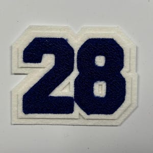 以下が含まれることがあります： 白のフェルトパッチに、ネイビーブルーで数字の28が描かれています。数字は白で縁取られ、レイヤー効果を生み出しています。パッチはクラシックでカレッジ風のスタイルです。