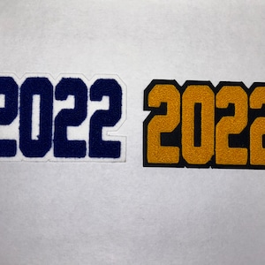 以下が含まれることがあります： 2022年が大きく白い文字で書かれたフェルトパッチ2枚。左のパッチは紺色、右のパッチは金色です。