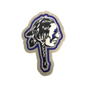May include: A blue and white embroidered patch of an indigenous person's head with a feather headdress. The patch is on a beige background.