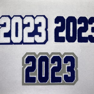 以下が含まれることがあります： 白い、紺色、そして灰色の輪郭のある紺色の3つのフェルトパッチに、2023という数字が書かれています。パッチは白い背景の上にあります。