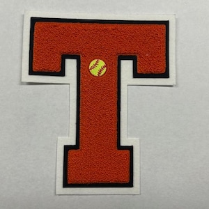 以下が含まれることがあります： 黒い輪郭と白い縁取りのある赤いフェルトの文字T。黒いステッチのある黄色のソフトボールが文字の上部に中央に配置されています。