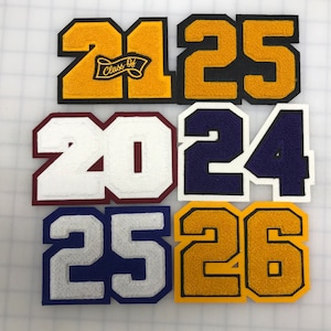 以下が含まれることがあります： 2125、2024、2526の数字がデザインされた、カラフルなフェルトパッチの詰め合わせ。パッチは、ゴールド、ブラック、ホワイト、ネイビーブルー、マルーンなど、さまざまな色があります。2125のパッチには「Class Of」の文字が含まれています。